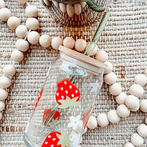 May include: A clear glass jar with a bamboo lid and a green glass straw. The jar has a red and white strawberry design. The jar is sitting on a woven mat with wooden beads.