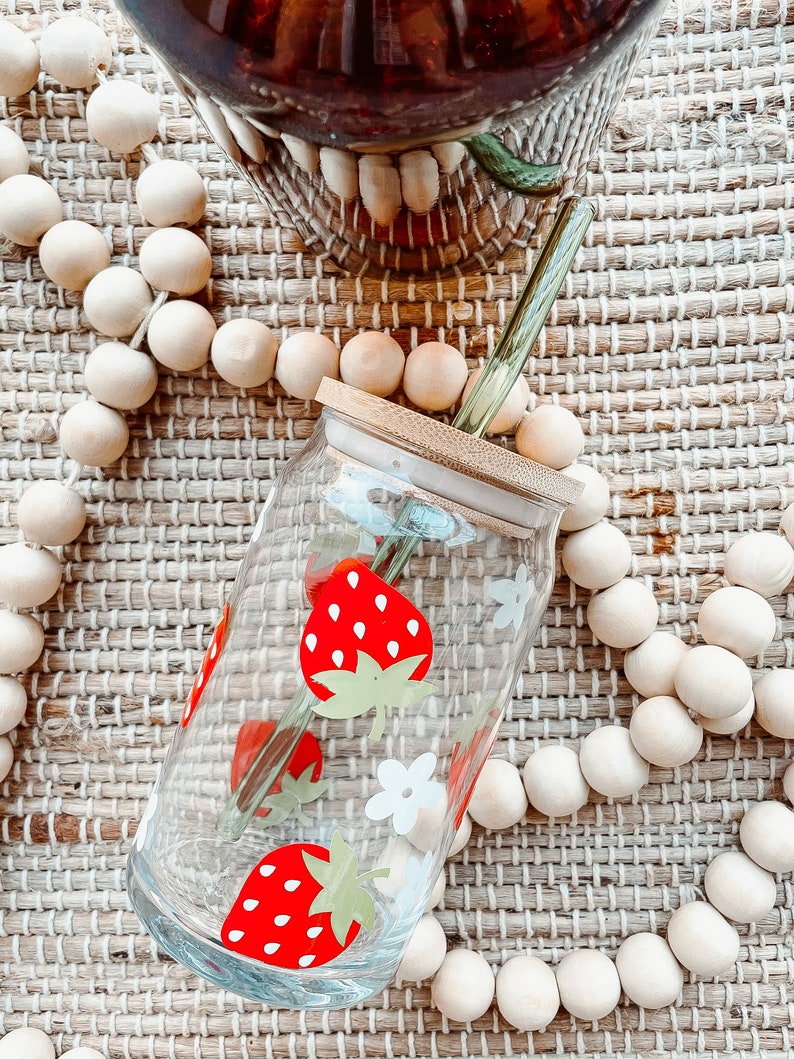 May include: A clear glass jar with a bamboo lid and a green glass straw. The jar has a red and white strawberry design. The jar is sitting on a woven mat with wooden beads.