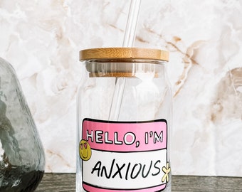 May include: A clear glass jar with a bamboo lid and a glass straw. The jar has a pink and black sticker that says "Hello, I'm Anxious" with a smiley face and a flower.
