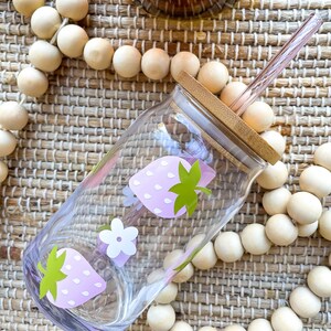 May include: A clear glass water bottle with a pink and green strawberry design. The bottle has a bamboo lid and a pink glass straw.