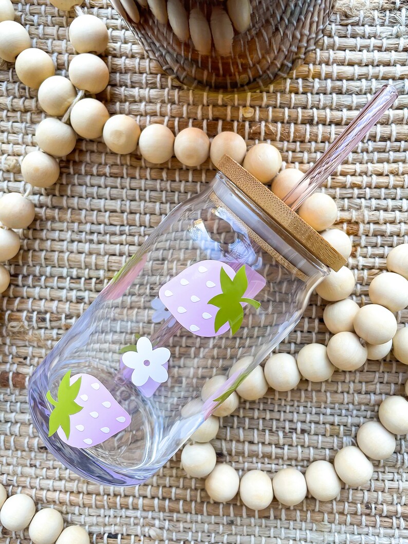 May include: A clear glass water bottle with a pink and green strawberry design. The bottle has a bamboo lid and a pink glass straw.