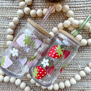 May include: Two clear glass drinking tumblers with bamboo lids and straws. One tumbler has a pink strawberry design and the other has a red strawberry design. Both tumblers are surrounded by wooden beads.
