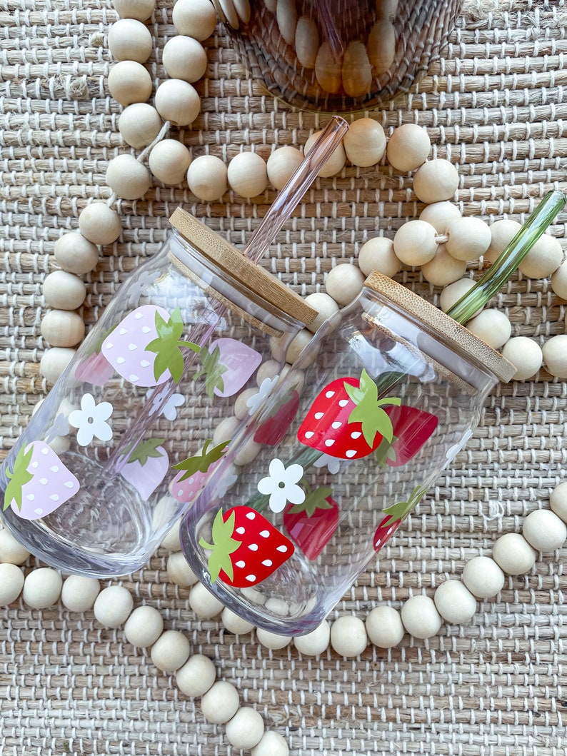May include: Two clear glass drinking tumblers with bamboo lids and straws. One tumbler has a pink strawberry design and the other has a red strawberry design. Both tumblers are surrounded by wooden beads.