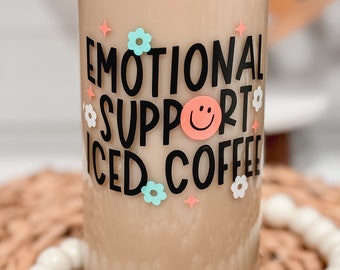 May include: A glass can with a light brown liquid inside. The can has a black and white design that says "Emotional Support Iced Coffee" with a smiley face and flowers.
