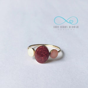 May include: Gold ring with three oval-shaped stones. The centre stone is a deep red, the stone on the left is white, and the stone on the right is pink.