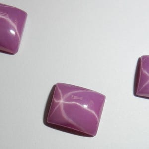 Könnte beinhalten: Drei rosa Stern-Saphire, jeder mit einem sechszackigen Sternmuster, werden auf einer weißen Oberfläche präsentiert. Die Saphire sind in rechteckiger Form geschliffen.