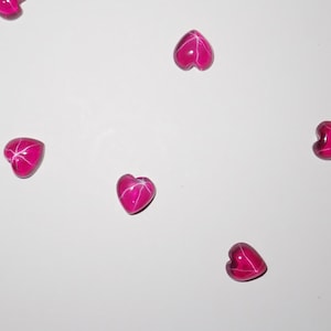 May include: A collection of bright pink heart-shaped beads scattered on a white surface. Each bead has a star-like pattern across its surface, creating a striking contrast against the solid pink color. The beads are glossy and appear to be made of glass or a similar material.