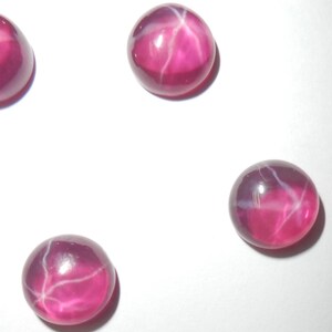 May include: Four round, polished gemstones with a star effect. The gems are a deep pink color with white star-shaped inclusions. The stones are set against a white background.
