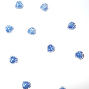 Puede incluir: Un conjunto de cuentas azules en forma de corazón con diseños blancos de estrella. Las cuentas están dispersas sobre un fondo blanco, creando un patrón visualmente atractivo. Adecuado para la creación de joyas u otros proyectos de manualidades.