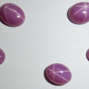 May include: Five pink star sapphires with a six-pointed star pattern. The sapphires are oval shaped and have a smooth, polished surface.