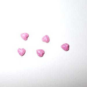 May include: Six pink heart-shaped gemstones are scattered on a white surface. Each gem has a star-like pattern on its surface. The gems are a vibrant pink colour and appear to be polished.