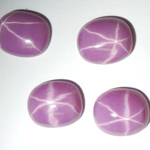 Könnte beinhalten: Vier rosa Sternrubin-Cabochons mit einem Sternmuster. Die Cabochons sind oval geformt und haben eine glatte, polierte Oberfläche.