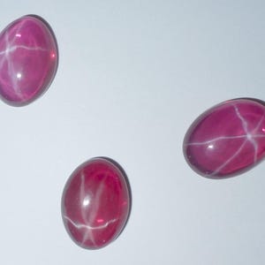 以下が含まれることがあります： 白い星模様の入ったピンクのスタールビーのカボションが3つ。カボションは楕円形で、光沢のある仕上げです。
