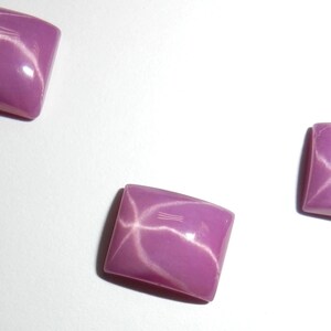 May include: Three rectangular cabochons with a smooth, polished surface in a vibrant lavender-purple hue. Each piece displays a distinct, centered star-like optical phenomenon with intersecting white light rays across the curved top surface.