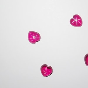 May include: Five pink heart-shaped stones with a starburst pattern inside. The stones are on a white background.