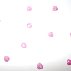 Puede incluir: Una dispersión de pequeñas cuentas rosas en forma de corazón sobre un fondo blanco. Cada cuenta tiene un acabado brillante y un sutil patrón de vetas blancas, creando una estética delicada y romántica. Los corazones están espaciados uniformemente.