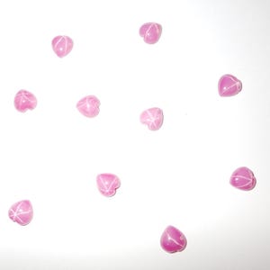 Puede incluir: Once cuentas rosas en forma de corazón con patrones de estrella blancos sobre un fondo blanco. Estas cuentas, adecuadas para la fabricación de joyas o manualidades, se presentan en una disposición visualmente atractiva. Cada cuenta tiene un diseño único de estrella.