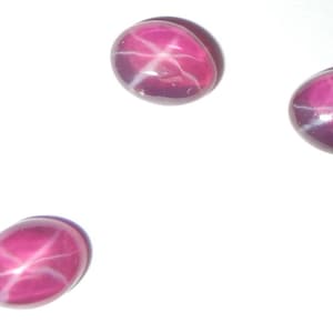 May include: Three pink star ruby cabochons with a white star in the center. The cabochons are oval shaped and have a smooth, polished surface.