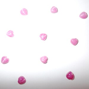 Puede incluir: Una colección de gemas rosas en forma de corazón dispersas sobre un fondo blanco. Cada gema tiene un efecto de estrella en el centro. Las gemas varían ligeramente en forma y tono de rosa.