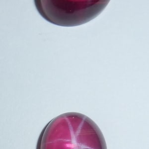 May include: Two oval-shaped, pink star ruby gemstones with a white star in the center.