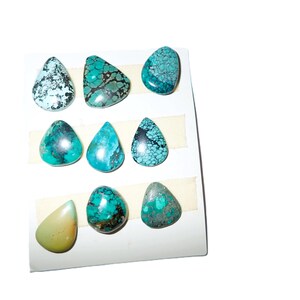 Può includere: Una collezione di otto cabochon di turchese lucidati disposti in tre file su un cartoncino bianco. Le pietre presentano varie tonalità di blu, verde e ottanio con motivi a matrice scura, inclusa una pietra verde chiaro liscia e tinta unita.
