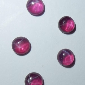 May include: Five pink star ruby cabochons with a white star in the center. The cabochons are round and have a smooth, polished surface.