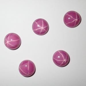 Pode incluir: Cinco pedras preciosas rosas, polidas e redondas, com um padrão de linhas brancas em forma de estrela. As gemas estão sobre um fundo branco, realçando a sua cor e o efeito de estrela. Cada gema tem cerca de 1 cm de diâmetro.