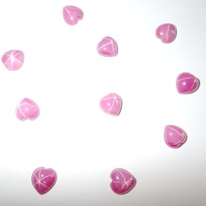 Puede incluir: Una colección de diez gemas rosas en forma de corazón con patrones blancos en forma de estrella. Las gemas están dispersas sobre un fondo blanco, mostrando sus superficies pulidas y diseños internos únicos. Adecuadas para joyería o manualidades.