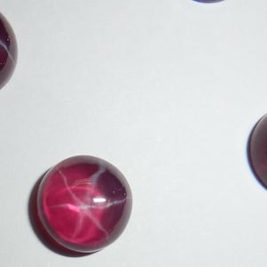 May include: Several cabochon-cut gemstones with a deep red hue and a star-like effect. The gems have a smooth, polished surface and a translucent appearance. The star effect is created by intersecting lines of light within the stone.