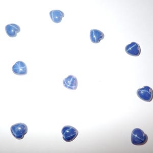 Puede incluir: Un conjunto de gemas azules en forma de corazón con efecto de estrella. Las gemas están dispersas sobre una superficie blanca, mostrando su apariencia pulida y patrones de estrella únicos. Ideales para la creación de joyas.
