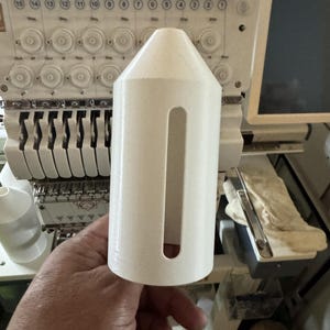 May include: A white, cylindrical object with a rectangular cutout, held in front of an embroidery machine. The object has a conical top and appears to be made of plastic. The background includes parts of the embroidery machine.