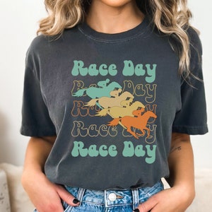May include: A dark gray t-shirt with a retro design featuring the text "Race Day" repeated four times with a different color horse graphic for each repetition. The horses are in shades of orange, yellow, green, and blue.