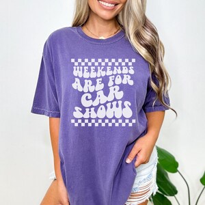 Pu&ograve; includere: Maglietta oversize viola con una grafica bianca che dice "Weekends are for car shows" in un font retr&ograve;. La grafica &egrave; circondata da un bordo a scacchi.