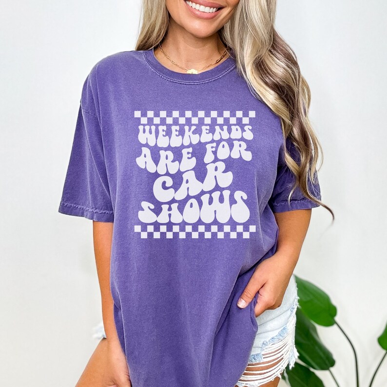 Pu&ograve; includere: Maglietta oversize viola con una grafica bianca che dice "Weekends are for car shows" in un font retr&ograve;. La grafica &egrave; circondata da un bordo a scacchi.