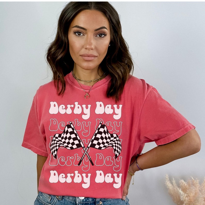 K&ouml;nnte beinhalten: Ein pinkfarbenes T-Shirt mit dem Text "Derby Day", der viermal in einer Retro-Schrift wiederholt wird. Der Text ist wei&szlig; umrandet. Zwei karierte Flaggen sind hinter dem Text gekreuzt.