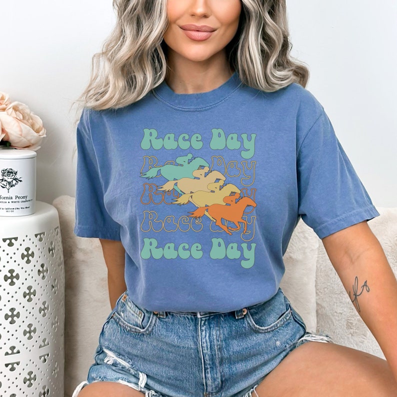 May include: A blue t-shirt with a retro design featuring the text "Race Day" repeated four times in a wavy font. The design also includes four stylized horses running in a line.
