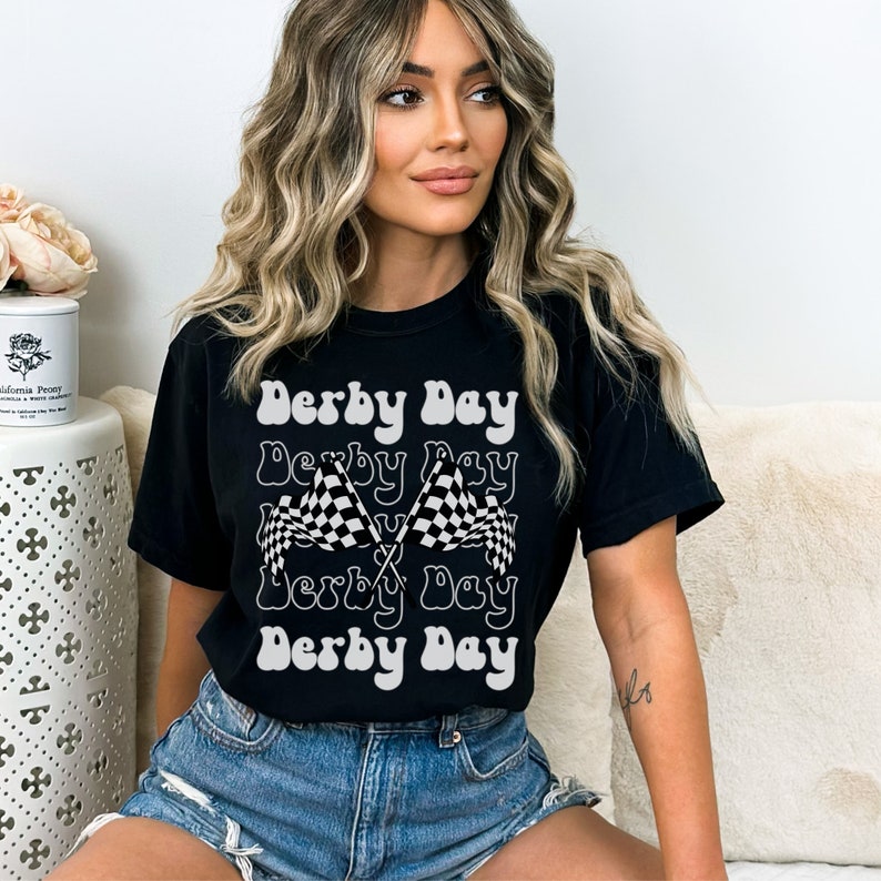 K&ouml;nnte beinhalten: Ein schwarzes T-Shirt mit einem wei&szlig;en Grafikdesign, auf dem in einer Retro-Schrift "Derby Day" steht. Die Grafik zeigt au&szlig;erdem zwei &uuml;berkreuzte karierte Flaggen.