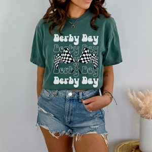 K&ouml;nnte beinhalten: Ein gr&uuml;nes T-Shirt mit dem Text "Derby Day", der in einer Retro-Schrift wiederholt wird. Das Shirt zeigt zwei gekreuzte karierte Rennflaggen.