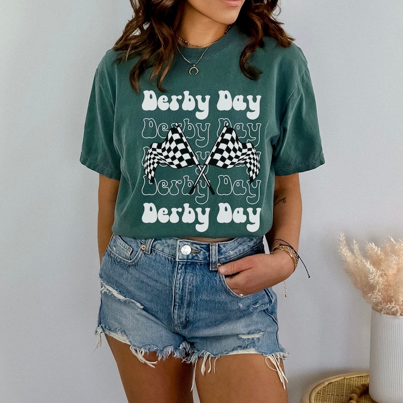 K&ouml;nnte beinhalten: Ein gr&uuml;nes T-Shirt mit dem Text "Derby Day", der in einer Retro-Schrift wiederholt wird. Das Shirt zeigt zwei gekreuzte karierte Rennflaggen.