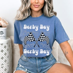 K&ouml;nnte beinhalten: Ein blaues T-Shirt mit dem Text "Derby Day" in einer Retro-Schrift. Der Text wird mehrmals wiederholt und ist &uuml;ber zwei karierte Rennflaggen gelegt.