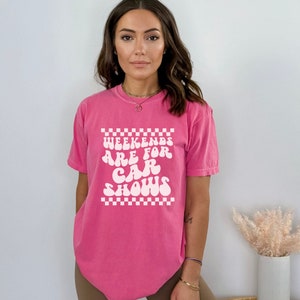 Pu&ograve; includere: Una maglietta rosa con una grafica bianca che dice "Weekends are for car shows" in un font retr&ograve;. La grafica &egrave; circondata da un motivo a scacchi bianco e nero.