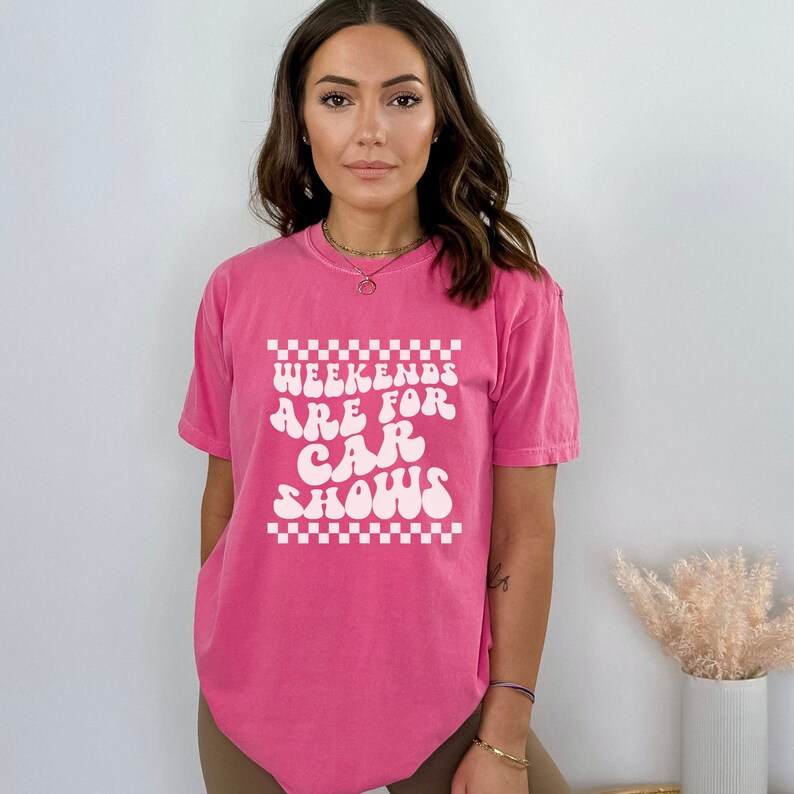 Pu&ograve; includere: Una maglietta rosa con una grafica bianca che dice "Weekends are for car shows" in un font retr&ograve;. La grafica &egrave; circondata da un motivo a scacchi bianco e nero.