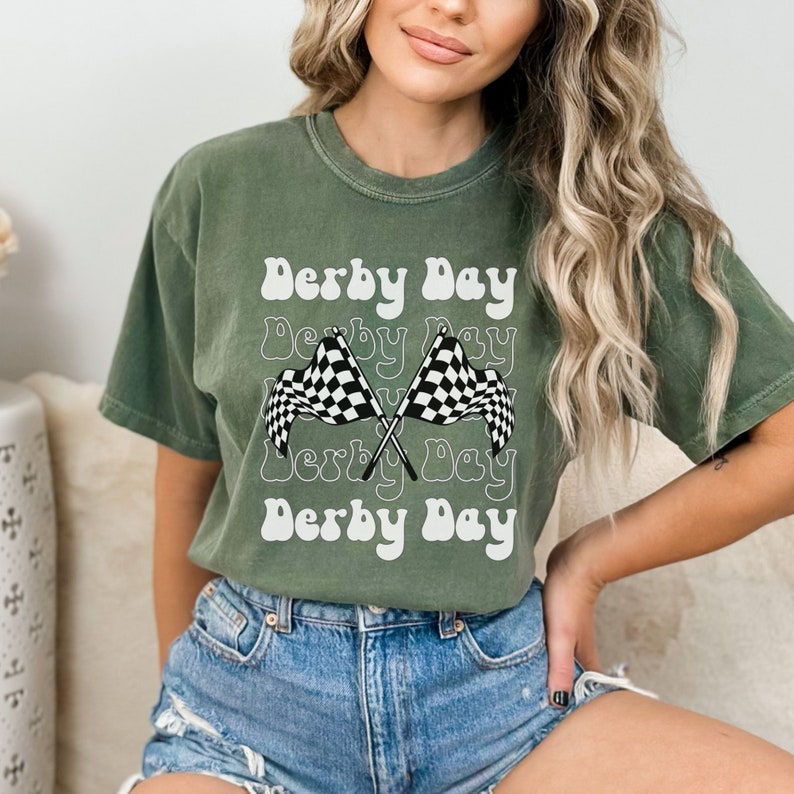 K&ouml;nnte beinhalten: Ein gr&uuml;nes T-Shirt mit dem Text "Derby Day", der in einer Retro-Schrift wiederholt wird. Zwei karierte Flaggen sind in der Mitte des Shirts gekreuzt.