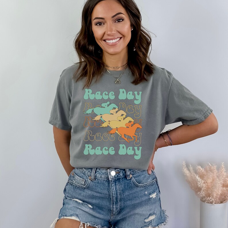 May include: A gray t-shirt with a retro-style graphic of horses racing. The graphic is in shades of orange, yellow, green, and blue. The text "Race Day" is repeated four times in a wavy font.