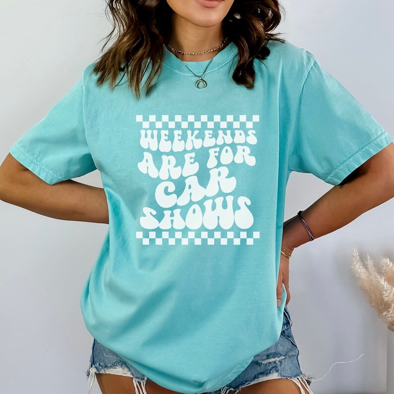 Pu&ograve; includere: Una maglietta blu turchese con una grafica bianca che dice "Weekends are for car shows" in un font retr&ograve;. La grafica &egrave; circondata da un bordo a scacchi.