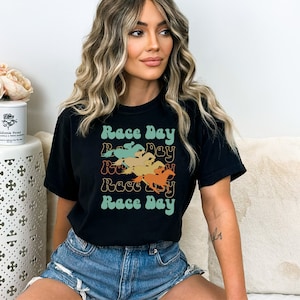 May include: Black t-shirt with a retro design featuring the text "Race Day" repeated four times in a stacked, layered design. Each layer of the text is a different color, with a silhouette of a horse and jockey in each layer.