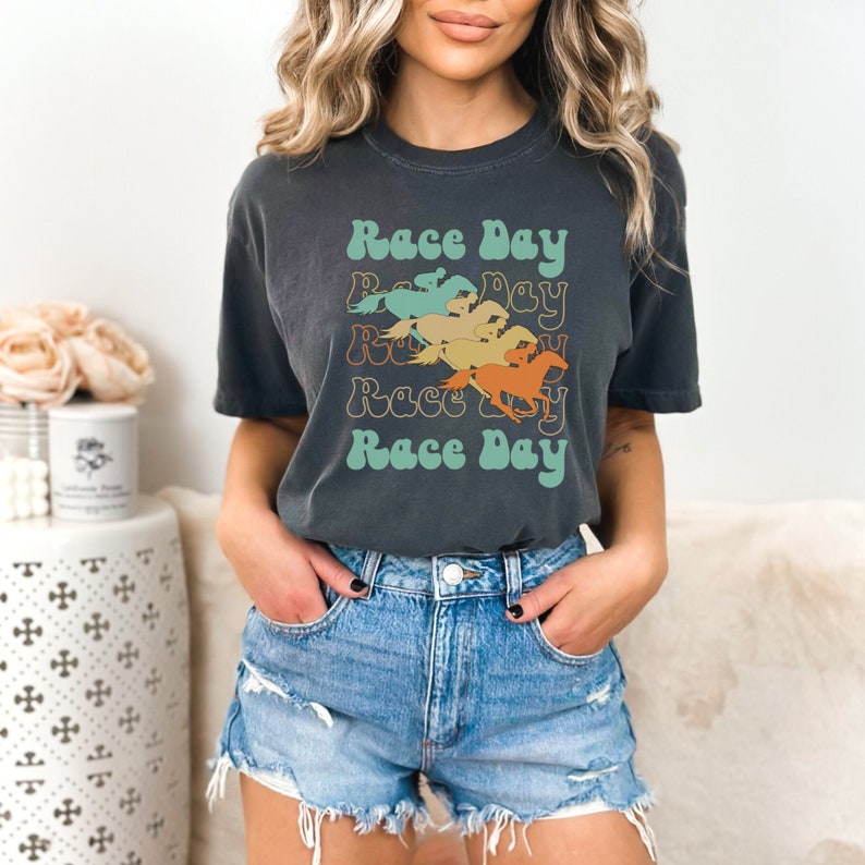 May include: A dark gray t-shirt with a retro design featuring the text "Race Day" repeated four times with a different color horse graphic for each repetition. The horses are in shades of orange, yellow, green, and blue.