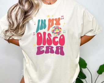 May include: A white t-shirt with the text "In My Disco Era" in colorful retro font. A disco ball graphic is included in the text.