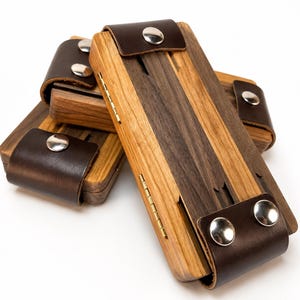 May include: A set of rectangular wooden cases featuring alternating light and dark wood grain panels. Each case is secured with a dark brown leather strap and silver-toned metal snap buttons, with visible brass hinges along the side edges.