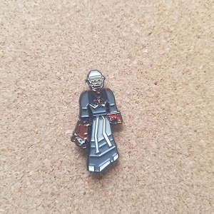 May include: A black and white enamel pin of a person wearing a white helmet, black clothing, and a red and white striped scarf. The person is holding a red and white object in their right hand.